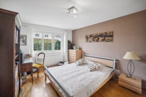 Une chambre bien organisée avec un lit double, un bureau, une chaise et une commode, avec du parquet et une fenêtre laissant passer la lumière naturelle.