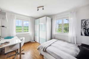 Chambre lumineuse avec un lit simple, une armoire et un bureau près de la fenêtre donnant sur les champs verdoyants.