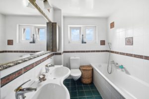 Une salle de bain lumineuse et propre avec une baignoire, des toilettes, un bidet, un lavabo et deux miroirs, avec des carreaux blancs avec des bordures décoratives et un sol bleu.