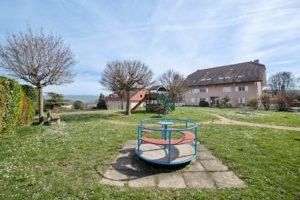 Un rond-point coloré dans une aire de jeux ensoleillée avec une structure de jeu en bois en arrière-plan, entouré de maisons d'habitation et d'un ciel clair.
