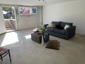 Un salon peu meublé avec un canapé gris, une petite table en osier, un tapis en peau d'ours et un sol en béton. une moto est visible à travers la porte ouverte.