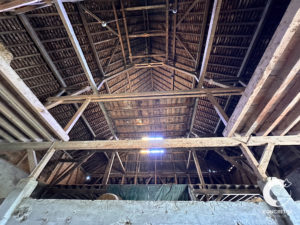 Vue intérieure d'une ancienne grange montrant une structure de toit en treillis de bois avec des panneaux manquants et une seule source de lumière.