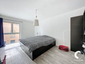 Une petite chambre moderne avec un lit double, un sol sombre, des murs blancs et une grande fenêtre avec des rideaux bleus.