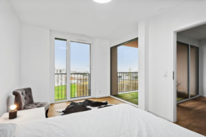 Chambre moderne avec un grand lit, des murs blancs, du parquet et deux portes ouvertes menant à un balcon avec vue sur une pelouse.