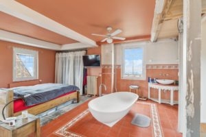 Chambre lumineuse et éclectique avec une baignoire îlot, des poutres apparentes, un grand lit et une télévision murale, avec un sol en terre cuite et un ventilateur de plafond.