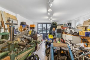 Intérieur d'un garage-atelier encombré rempli d'outils, de bois, d'une moto, de vélos et de modèles réduits de voitures près d'une fenêtre.