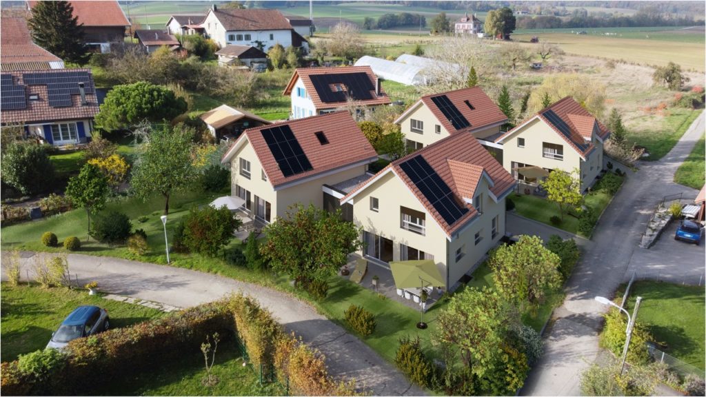 Vue aérienne d'un quartier résidentiel avec des maisons modernes équipées de panneaux solaires. Le paysage environnant comprend de la verdure, des routes et des arbres épars.