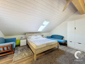 Chambre mansardée avec un lit en bois, deux canapés bleus, une petite table de nuit et une armoire blanche. Une fenêtre de toit se trouve sur le plafond en pente.