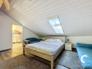 Une chambre mansardée confortable avec un plafond en pente, une lucarne et un lit double. La chambre dispose d'un canapé bleu, d'un fauteuil et d'un tapis. Une salle de bain est visible à travers une porte ouverte.