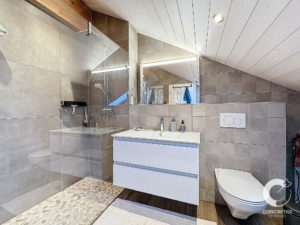 Salle de bain moderne avec plafond en pente comprenant une douche vitrée, un lavabo blanc avec vanité, des toilettes murales et des murs en carrelage neutre.