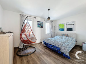 Une chambre moderne avec un lit, une chaise suspendue, deux fenêtres et des œuvres d'art murales. Un plancher en bois et un luminaire au plafond complètent la décoration de la pièce.