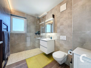 Salle de bain moderne avec de grands murs carrelés, une vanité flottante avec lavabo, une cabine de douche en verre et des toilettes murales. Une fenêtre laisse entrer la lumière naturelle. Les accessoires de bain sont visibles sur la vanité.