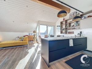 Séjour et cuisine modernes à aire ouverte avec îlot noir, canapé jaune, touches de bois et étagères décoratives. Les fenêtres au plafond offrent une abondante lumière naturelle.