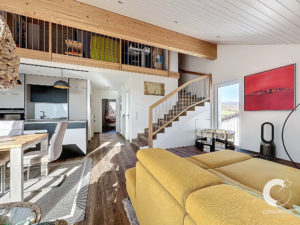 Espace de vie moderne à aménagement ouvert, comprenant un canapé jaune, un coin repas, une cuisine et un loft. Des escaliers en bois mènent au niveau supérieur. De grandes fenêtres et des œuvres d'art ornent les murs.