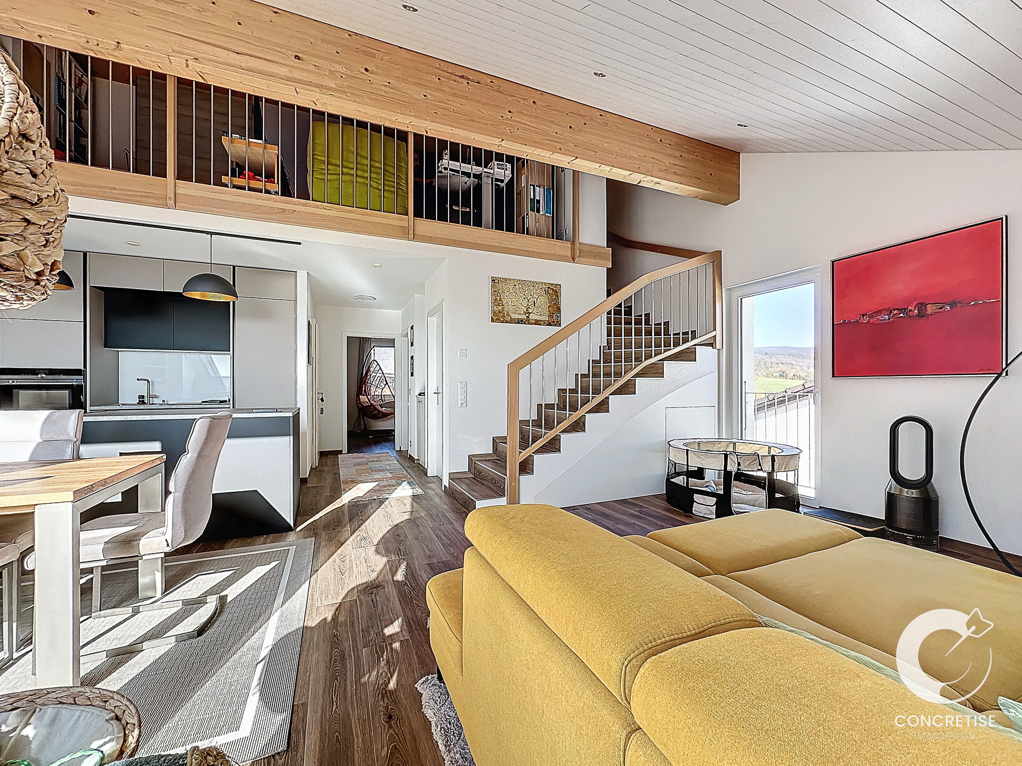 Espace de vie moderne à aménagement ouvert, comprenant un canapé jaune, un coin repas, une cuisine et un loft. Des escaliers en bois mènent au niveau supérieur. De grandes fenêtres et des œuvres d'art ornent les murs.