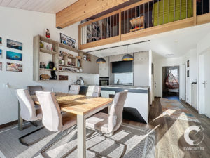 Salle à manger moderne avec table en bois et six chaises, adjacente à une cuisine aux armoires épurées. Au-dessus se trouve un espace mezzanine avec balustrade, et les murs sont ornés de photos et d'objets décoratifs.
