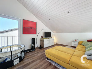 Chambre mansardée confortable avec un canapé d'angle jaune, un meuble TV et un parc pour chien. Un tableau rouge orne le mur et le plafond est incliné avec des panneaux en bois.