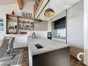 Cuisine moderne avec îlot gris, luminaires suspendus et étagères ouvertes exposant divers objets. Coin repas avec chaises rembourrées à gauche. Le plafond présente des poutres en bois apparentes.