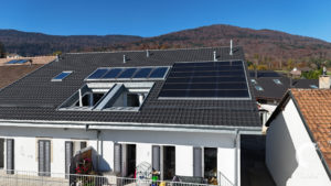 Une maison moderne avec des panneaux solaires et des puits de lumière sur le toit, sur fond de collines boisées sous un ciel bleu clair.