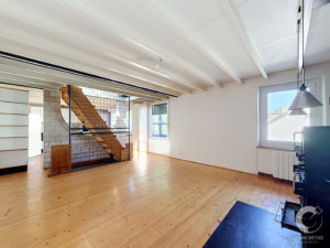 Open-plan loft with exposed white ceiling beams, wooden floor, and a metal-framed staircase leading to a mezzanine or upper level, near large windows.
