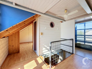 Indoor stairwell with white paneled ceiling, exposed wooden beams, blue accent wall, and metal railing beside a large blue-framed window.