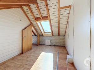 Attic room with a sloped ceiling, exposed wooden rafters, and a skylight window letting in daylight; a wooden door is ajar on the left edge of the frame.