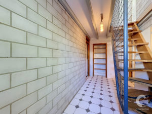 Narrow hallway with a pale green brick wall, white-and-gray tiled floor, and a wooden staircase with blue railing on the right.