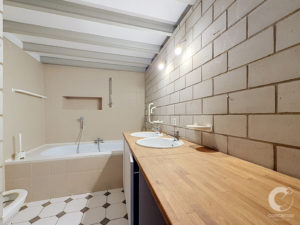 Bathroom with a long wooden counter and three sinks against a concrete block wall; tiled floor and a white bathtub to the left under a paneled ceiling.