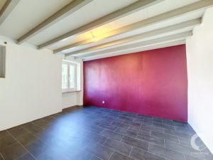 Empty room with a dark tile floor, white walls and exposed ceiling beams; a purple accent wall on the right and a window on the left letting in light.