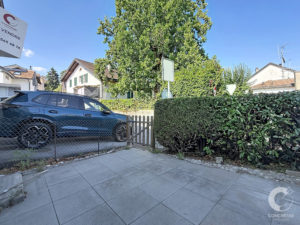 Residential street scene with a dark blue SUV behind a chain-link fence, a large leafy tree, and houses nearby in sunlight.