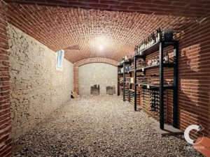 Underground wine cellar with brick arched ceiling, stone walls, and gravel floor; metal shelving holds rows of wine bottles.