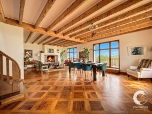 Open-plan living and dining room with exposed wooden beam ceiling, parquet floors, and a stone fireplace; a dark dining table with teal chairs sits near large windows.