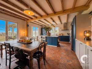Open-plan kitchen and dining area with blue cabinets, exposed wooden beams, and a large wooden dining table topped with a fruit bowl.