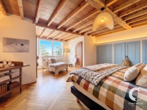 Cozy bedroom with exposed wooden beams, a plaid quilt bed, and a seating area by a sunny window. Warm lighting fills the space.