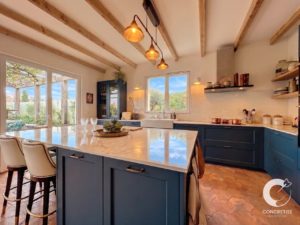 Bright kitchen with a blue island, marble countertop, and copper pendant lights overlooking a garden through glass doors.