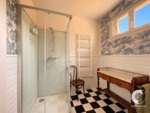 Bathroom with a glass corner shower, white tiled walls, and a wooden marble-top vanity against a blue tree-pattern wallpaper.