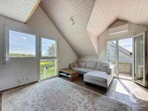 Bright living room with a white sectional sofa, wooden coffee table, and a patterned rug under sloped white-paneled ceilings; large windows overlook a green lawn and blue sky.