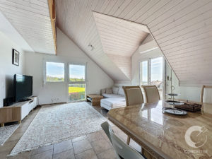 Attic living room with a sloped white wooden ceiling, large windows, and a marble dining table in the foreground.