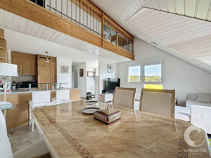 Open-plan loft with a marble dining table, beige chairs, and a wood kitchen island beneath a white paneled ceiling.
