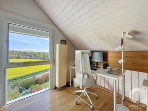 Bright workspace by a large window with a white desk, ergonomic chair, monitor, and floor lamp against a slanted wooden ceiling.