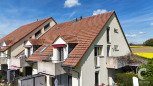 Row of cream-colored townhouses with red-tiled, sloped roofs under a blue sky; chimneys and balconies visible in a sunny countryside setting
