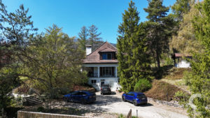 Two-story house with a red-tiled roof surrounded by tall trees and a driveway with several cars under a bright blue sky.