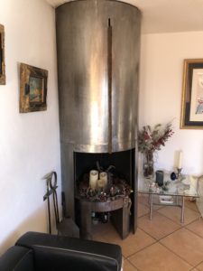 Tall metallic cylindrical fireplace in a living room, with Christmas decorations and framed paintings on the walls.