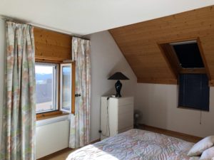 Bedroom with a slanted wooden ceiling, two single beds in pastel floral quilts, and a white dresser under a window with patterned curtains exterior light offers a view.