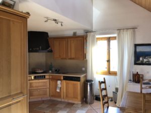 Kitchen with light wood cabinets, a gas stove, and a window with white curtains letting in sunlight.