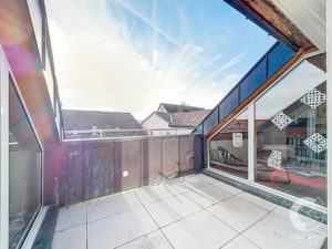 Sunny rooftop balcony with white tiled floor, dark panel railing, and large slanted glass façade; neighboring houses in the background.
