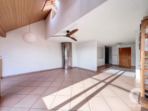 Bright, empty living room with a high wooden vaulted ceiling, skylights, and a ceiling fan; terracotta floor tiles and a metal chimney column. Large windows cast long shadows across the floor.