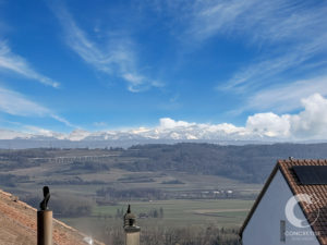 Snow-capped mountains under a clear blue sky, viewed from a village with red-tiled roofs in the foreground.