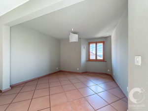 Empty interior room with pale blue walls, a brown tiled floor, and a wooden-framed window bringing in daylight.