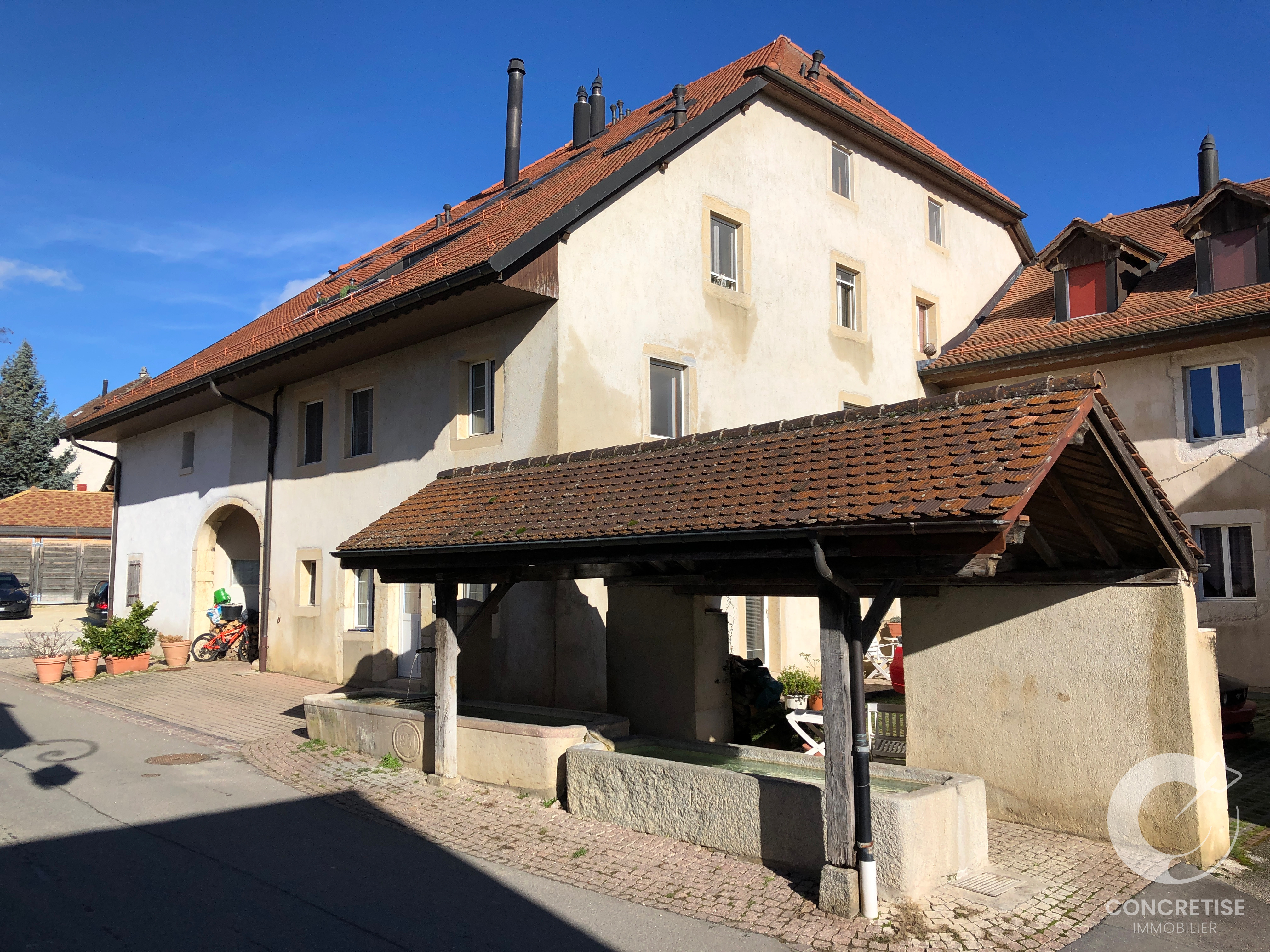 Two-story beige stucco house with a red-tiled roof and arched doorway, fronted by a covered stone trough and potted plants.